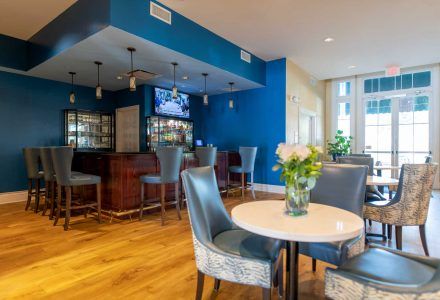 Bar with blue walls, wooden bar, stools, and tables with chairs; flowers on table, exit doors visible.