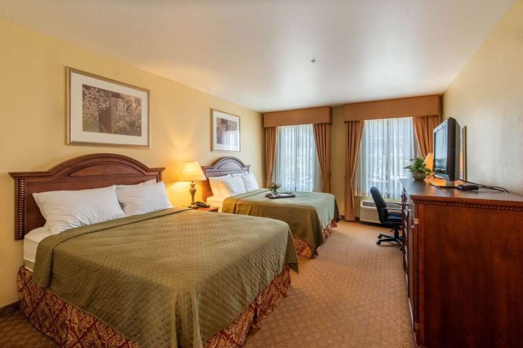 Hotel room with two queen beds, TV, desk, and curtains. Beige walls, brown furniture, tan carpet.