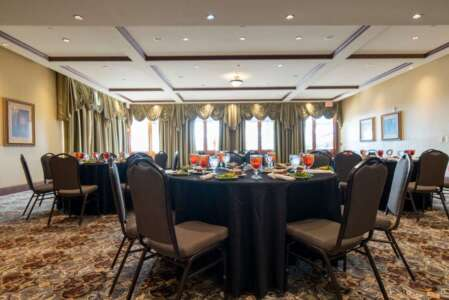 Round tables in a banquet room, set with place settings, dark tablecloths and chairs, windows with curtains.