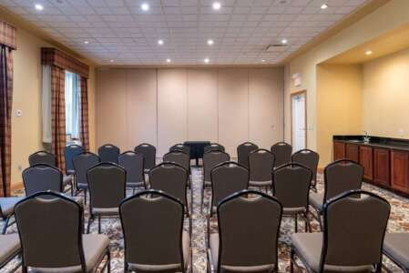 Rows of chairs face the front of a conference room with a beige wall, small table, and a wet bar.